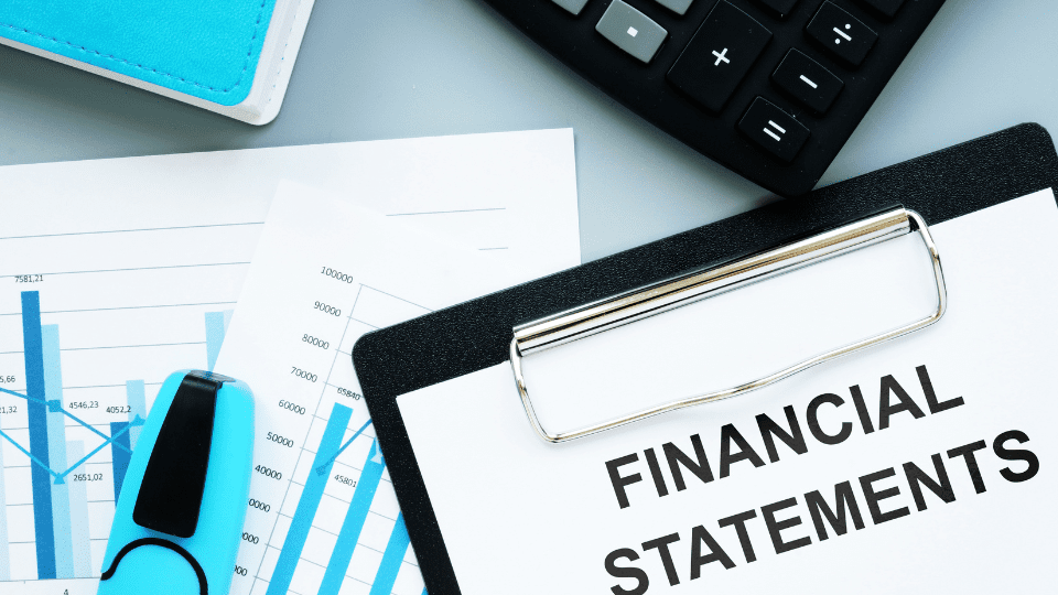 The words financial statement written on paper on a clipboard with a calculator, maker, and various financial graphs and figures around it 