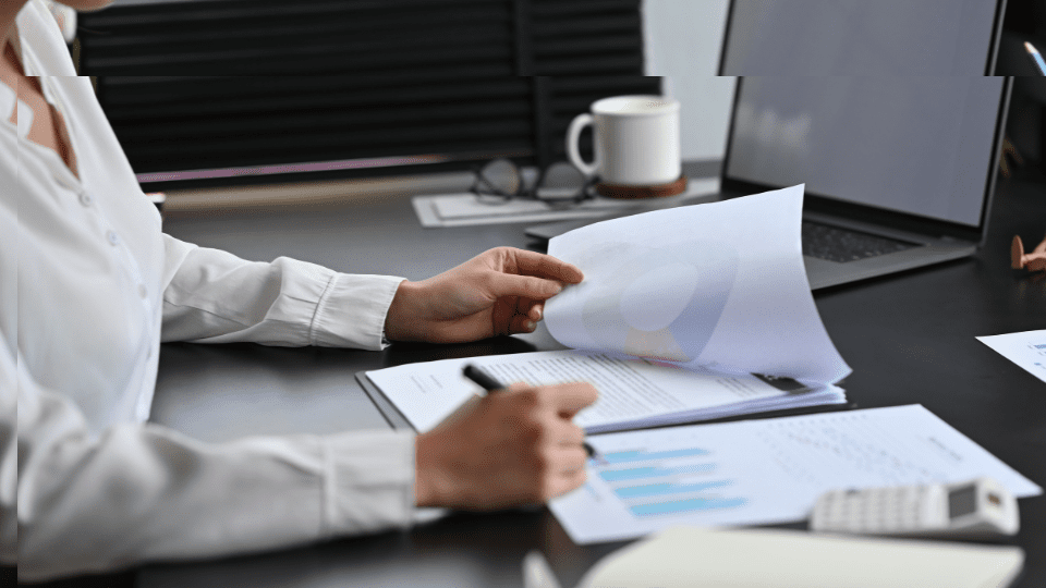 A person sitting at a desk preparing a report, looking at graphs, and holding a pen